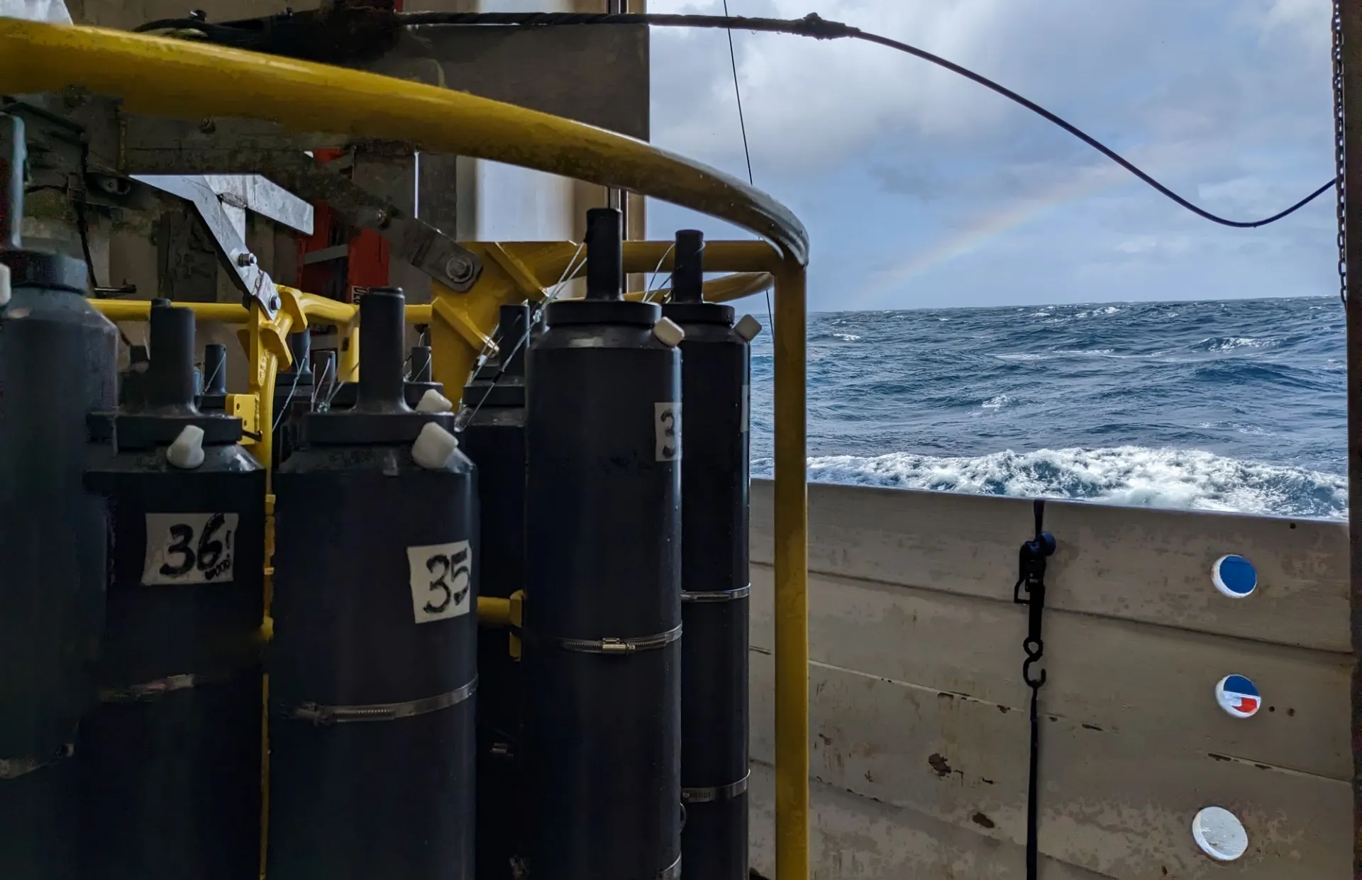 Photo of the Pacific Southern Ocean with seawater sampling bottles in foreground and faint rainbow in background, taken on GEOTRACES Cruise GP17-OCE